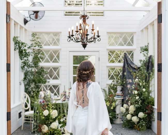 A woman in a white wedding dress stands at the entrance of a garden room, holding a bouquet and facing a beautifully decorated table surrounded by floral arrangements.