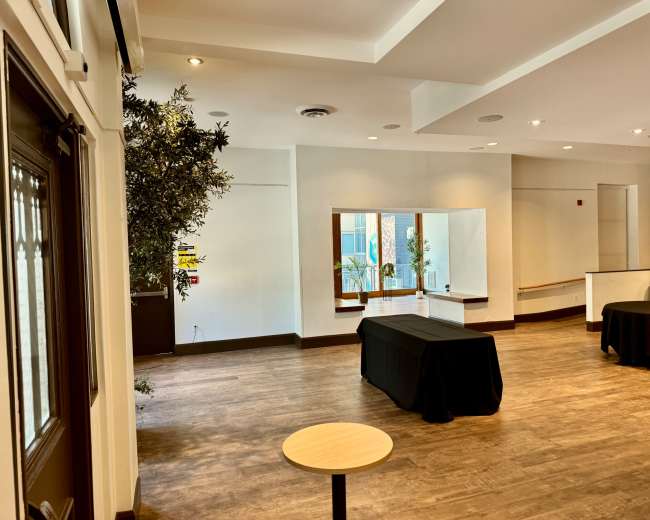 The image shows a spacious indoor area with wooden flooring, two black-covered tables, and a potted plant near a large window.