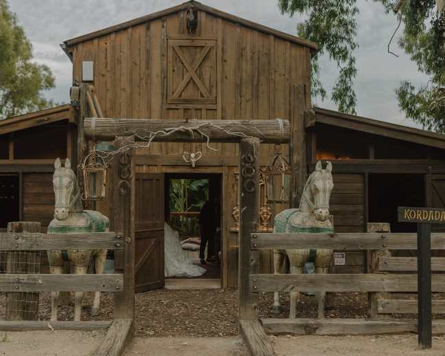 A rustic wooden barn features two carved horse statues at its entrance, flanked by a fenced walkway.