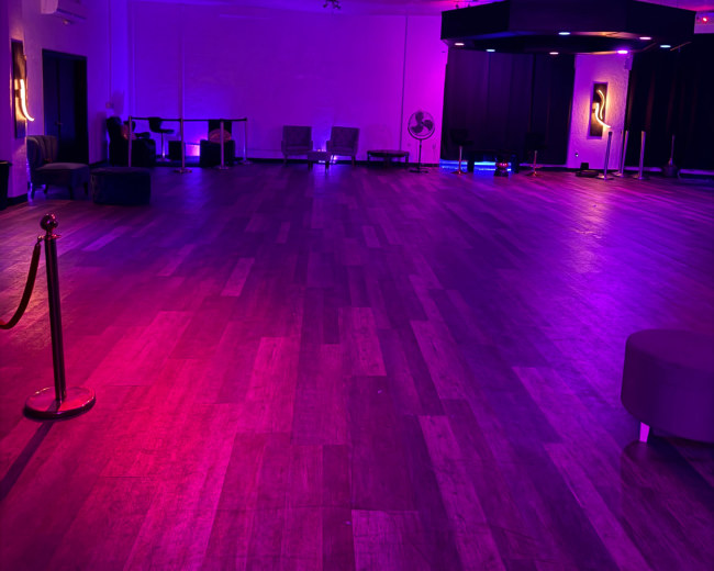 The image shows an empty event space with wooden flooring, illuminated by purple and pink lighting.