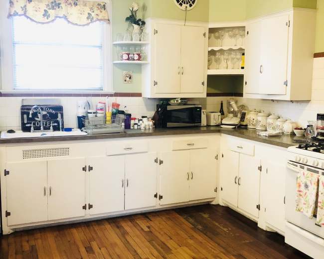 The image shows a kitchen with white cabinetry, a window adorned with floral curtains, and wood flooring.