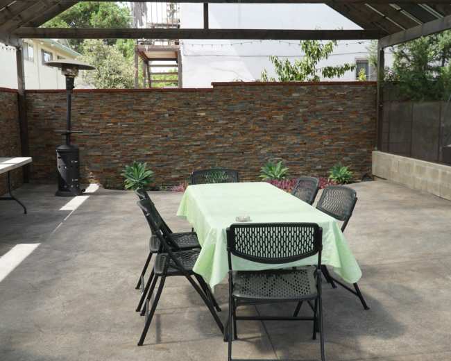 An outdoor patio features a table covered with a green tablecloth surrounded by black chairs, with a stone wall in the background.