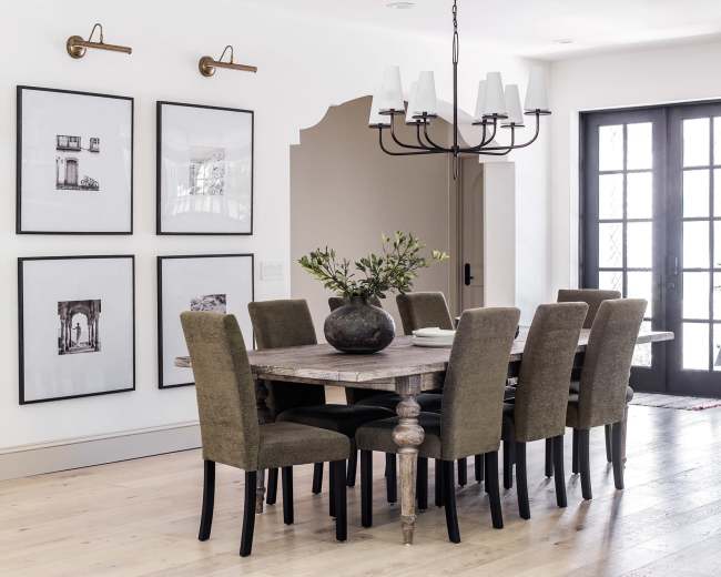 A dining room with a large wooden table surrounded by eight upholstered chairs, complemented by framed black-and-white photographs on the walls and a modern chandelier overhead.