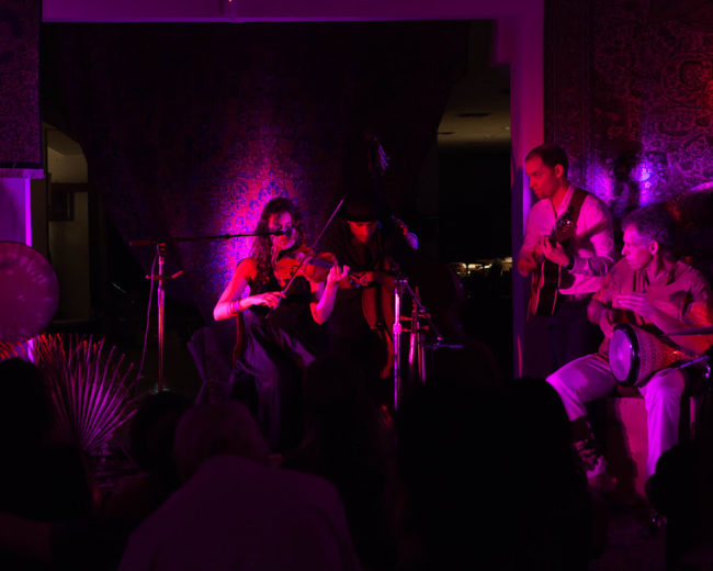 A group of musicians performs together on stage, illuminated by purple lights, with various instruments including a violin, guitar, and percussion.