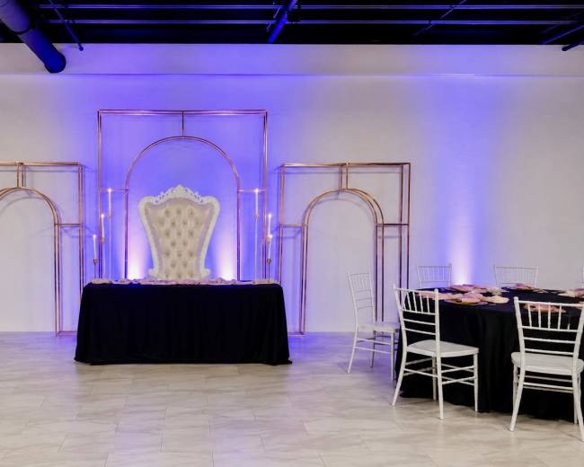 A lavish event space features a grand white throne center stage, flanked by gold arch decorations and illuminated with purple lighting, surrounded by elegantly set tables.