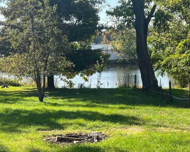 A fire pit is located in a grassy area with a view of a lake and trees in the background.