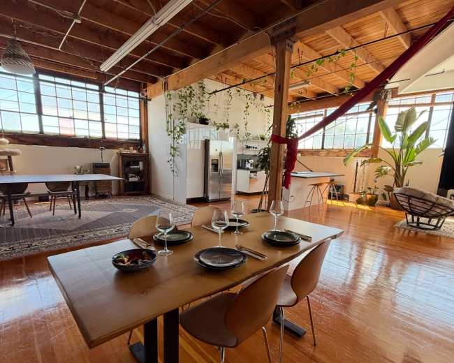 The image shows a spacious dining area with a wooden table set for four, surrounded by large windows and plants in a well-lit open space.