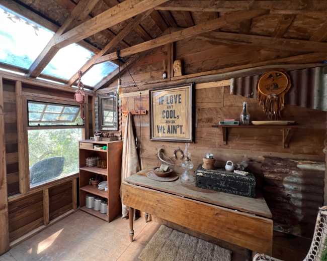 The interior of a rustic cabin features wooden walls, a table with various objects, and a large window allowing natural light to enter.