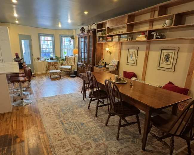 The image shows a spacious living area with a wooden dining table, chairs, and a cozy seating arrangement, illuminated by natural light from large windows.