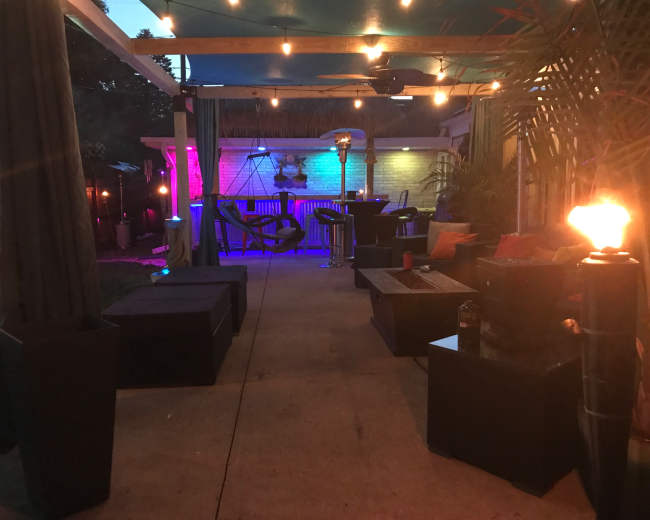 A backyard patio area illuminated by string lights, featuring seating arrangements with cushions, a table, and decorative elements like torches and colorful lighting.