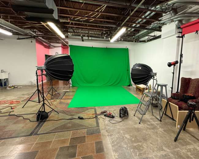 The image shows a photography studio setup featuring green backdrop, two large studio lights, and a seating area with two chairs.