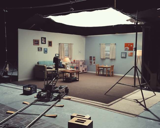 A film set features a cozy living room scene with a sofa, two chairs, children's toys, and a table arranged on a carpeted floor.