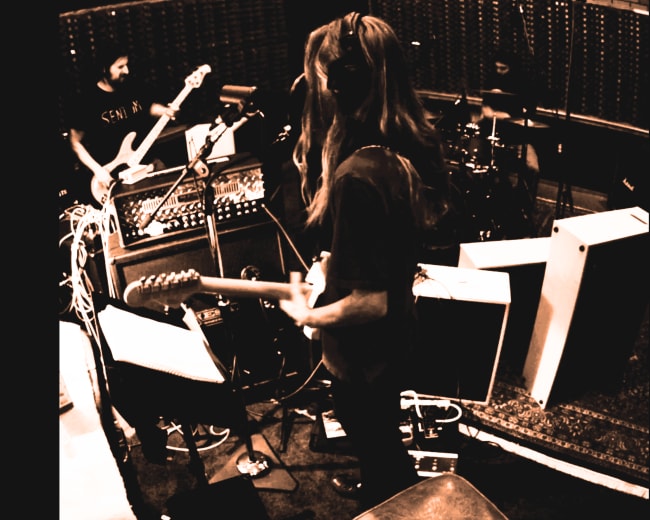 Musicians are recording in a studio with guitars, drums, and various equipment set up around them.