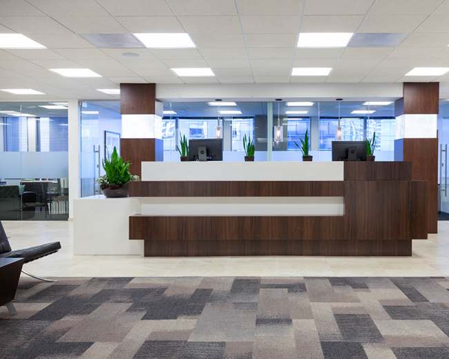 The image shows a modern reception area with a wooden front desk and potted plants, flanked by glass-enclosed office spaces.