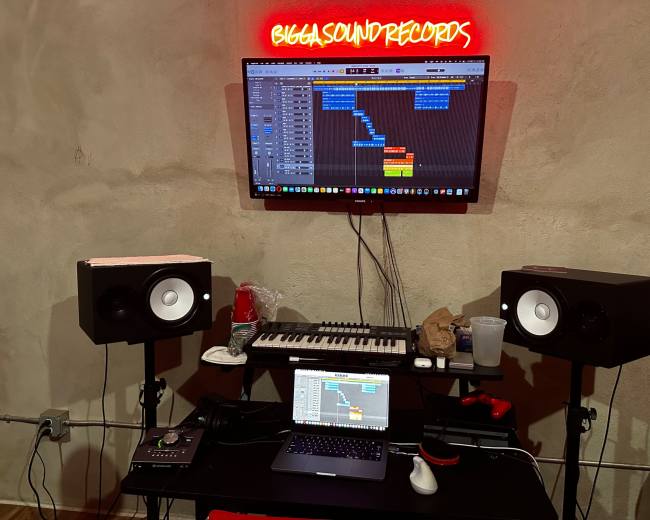 The image shows a music production setup with a computer screen displaying a digital audio workstation, two studio monitors, a keyboard, and various equipment on a dark table under a brightly lit sign.