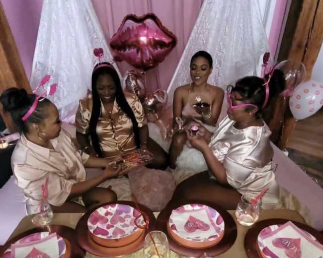 Four women gather around a decorated table with pink-themed treats and drinks in a festive setting.
