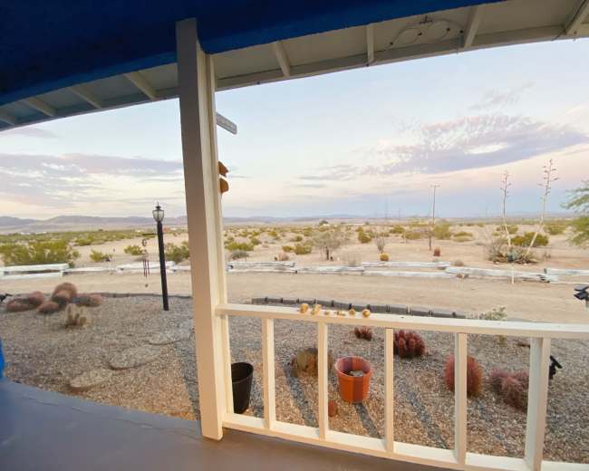 The image shows a view from a porch overlooking a desert landscape with sparse vegetation and a distant mountain range.