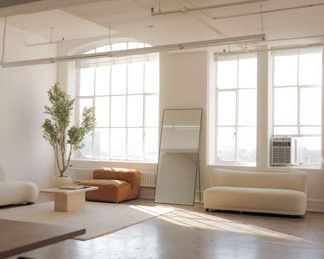 A bright, minimalist living room features large windows, a potted plant, an orange chair, a mirror, and two light-colored sofas.