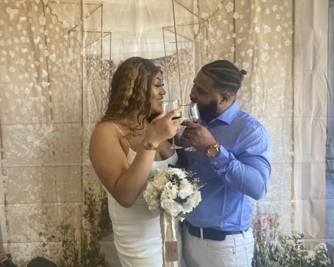 A couple shares a toast while holding glasses in front of a floral backdrop, with one partner holding a bouquet.