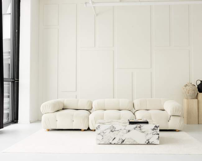 A light-colored, modern living room features a plush sectional sofa, a marble coffee table, and minimalistic decor against a neutral wall.