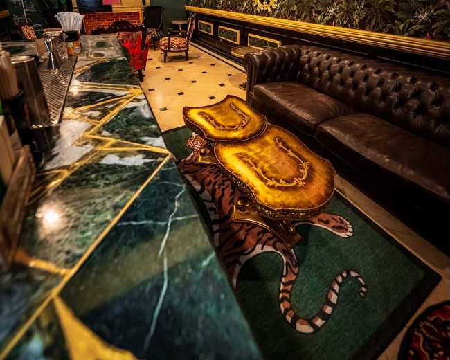 The image shows a stylish bar area featuring a dark leather sofa, a marble-topped counter, lush wallpaper, and unique decorative elements.