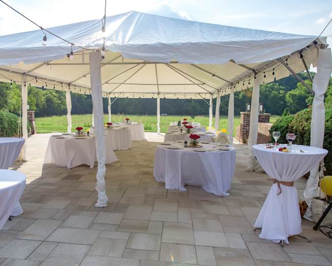 A large white tent with several round tables set for a gathering sits on a stone patio overlooking a green field.