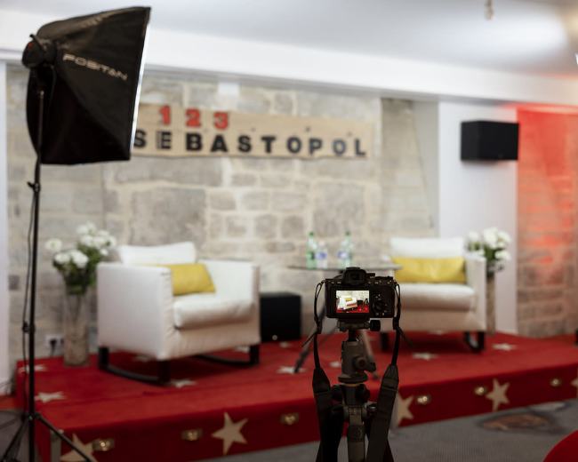 A camera is positioned on a tripod facing two white chairs with yellow cushions, set against a stone wall with the word "SEBASTOPOL" displayed above.