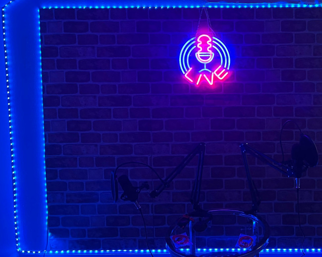 The image shows a podcast studio with a neon microphone sign and blue LED lights illuminating a brick wall, along with two microphone stands and a glass table.