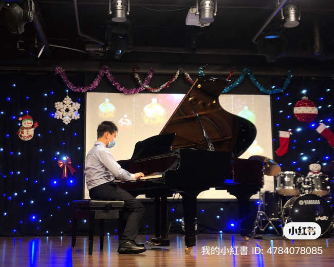 A person in a mask plays a grand piano on stage with decorative lights and a festive backdrop.
