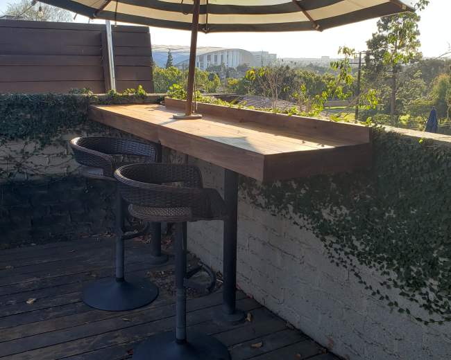A rooftop outdoor seating area with a wooden table and two wicker chairs, surrounded by greenery and an umbrella for shade.