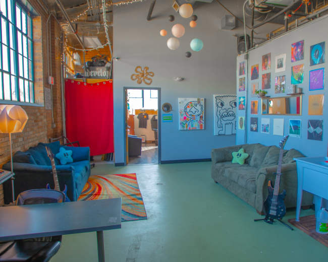 The image shows a colorful, decorated room with a seating area, artwork on the walls, and various decor items, including a guitar and a rug.