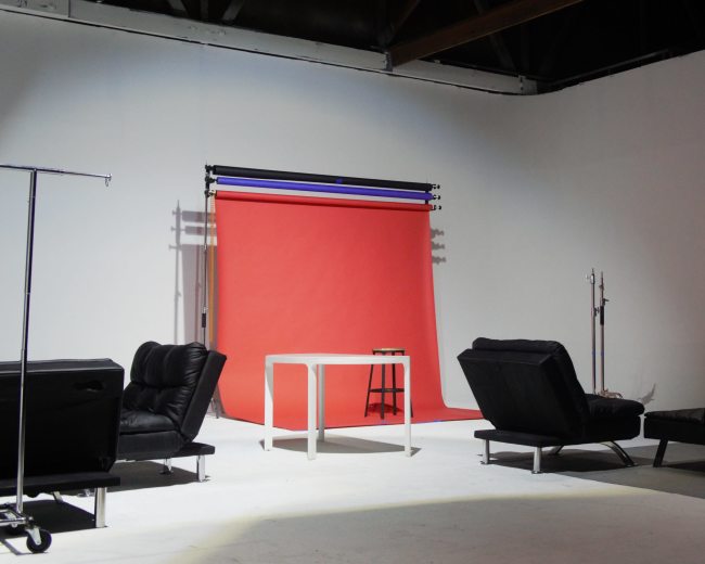 A photo studio features a red backdrop with a table and two black couches arranged around a minimalist space.