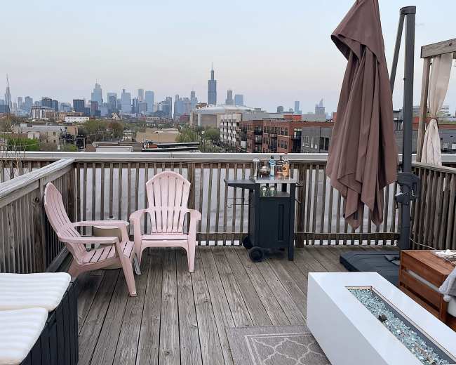 A rooftop patio features wooden decking, two pink chairs, a table with a fire pit, and a view of the Chicago skyline in the background.