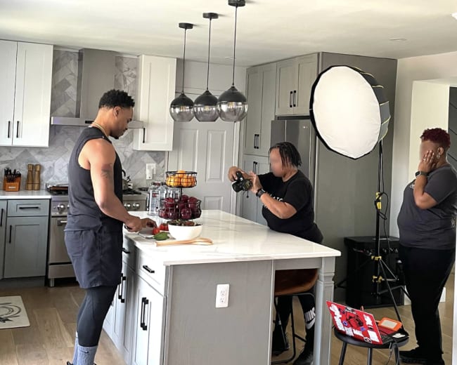 A man prepares food at a kitchen island while two photographers set up equipment to capture the scene.