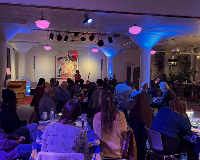 A performer stands on a small stage in a softly lit room filled with seated audience members, some at tables with drinks, while colorful lights illuminate the space.