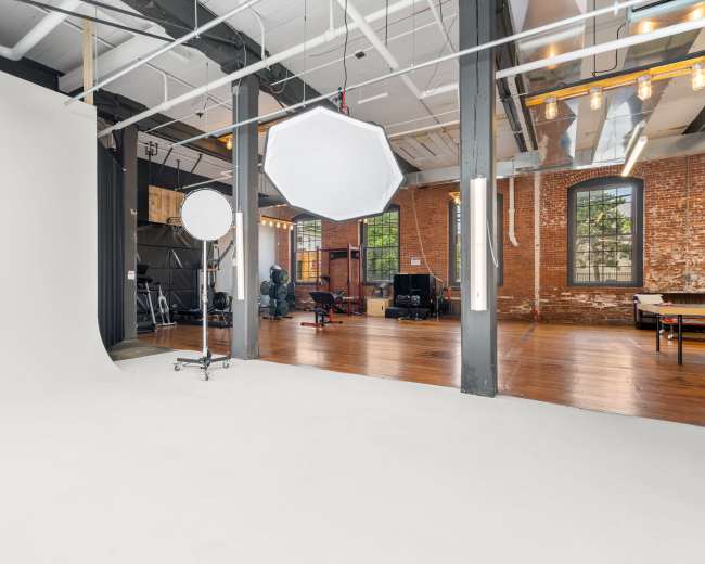 A spacious photography studio with a white backdrop, a large light modifier, and exposed brick walls, showcasing wooden flooring and various equipment in the background.