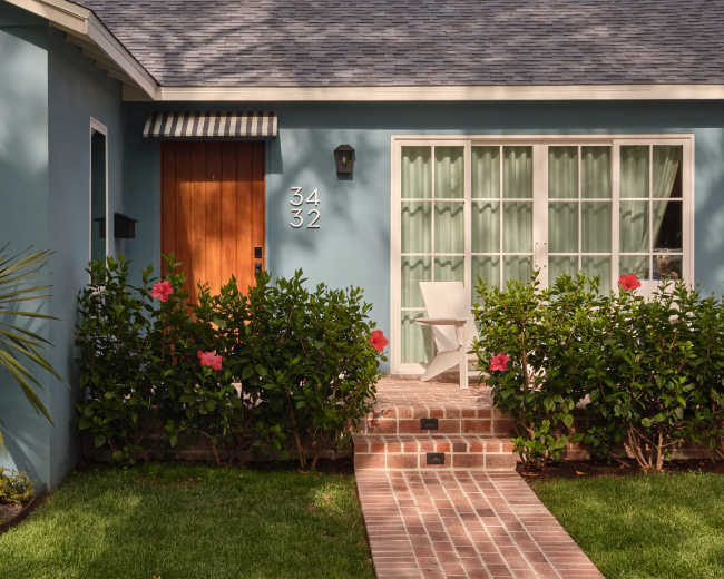 The image shows a well-maintained home facade with a blue exterior, a brick pathway, and green shrubbery, featuring a wooden door and a large window with sheer curtains.