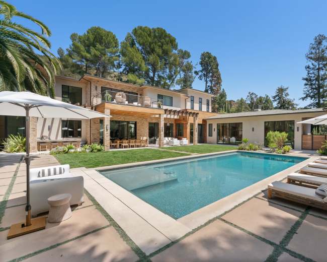 The image features a modern house with a spacious pool area surrounded by lounge chairs and landscaped greenery.