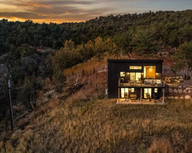 A modern two-story black house stands on a hillside surrounded by trees and rocky terrain at sunset.