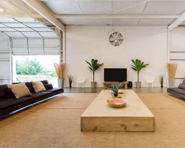 The image shows a spacious indoor area with two sofas facing each other, a large wooden coffee table in the center, a television on a stand, and potted plants adorning the space.