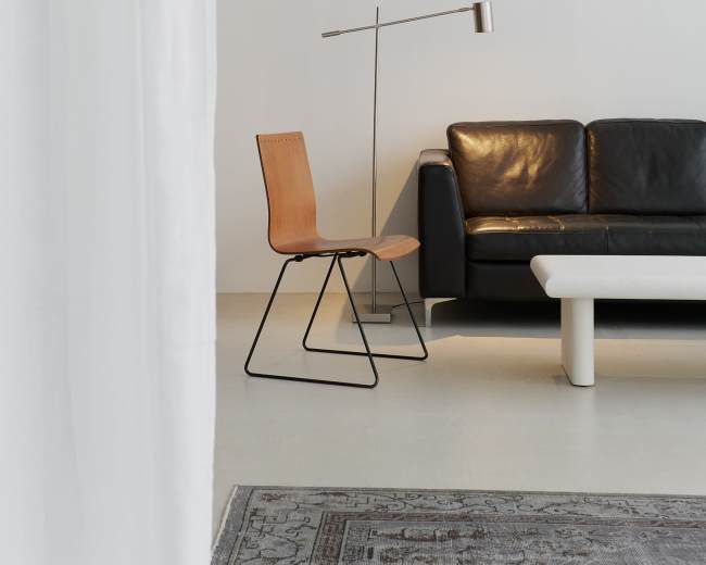 A minimalist interior features a black leather sofa, a light-colored coffee table, a wooden chair, and a modern floor lamp against a white wall.