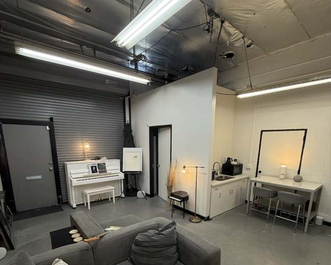 The image shows a spacious interior with a gray sectional sofa, a white piano, a small kitchen area, and a workspace with a table and chairs, all under a ceiling with exposed pipes and industrial lighting.