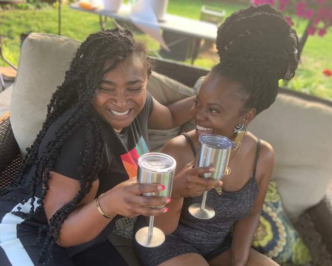 Two women are sitting on a patio couch, clinking stainless steel cups in celebration.