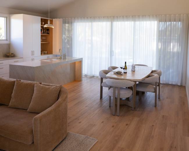 A spacious, modern interior features a kitchen island and a dining table surrounded by chairs, with large windows covered by sheer curtains allowing natural light to enter.