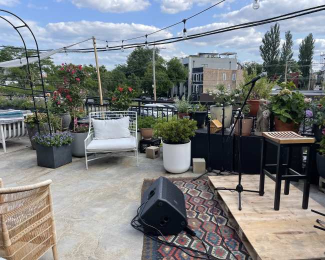 A rooftop space features potted plants, a microphone setup on a wooden platform, and seating arrangements with a scenic backdrop.