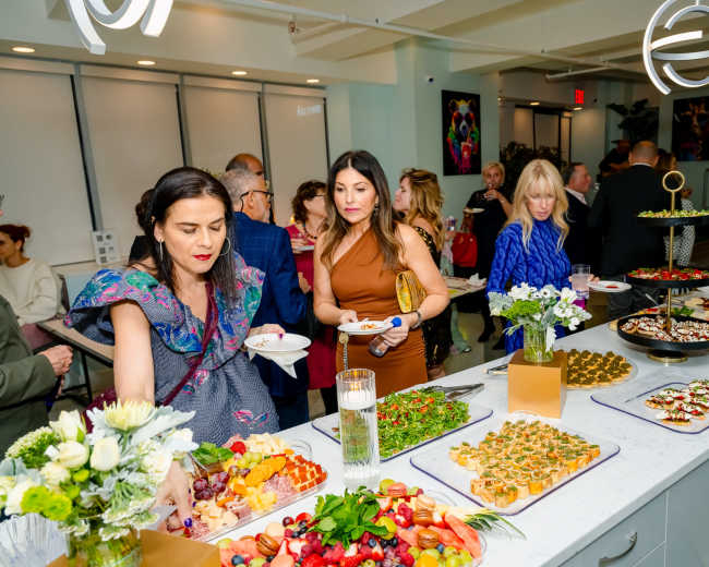 A group of people mingles at a well-decorated event with a variety of colorful food displays on a social table.