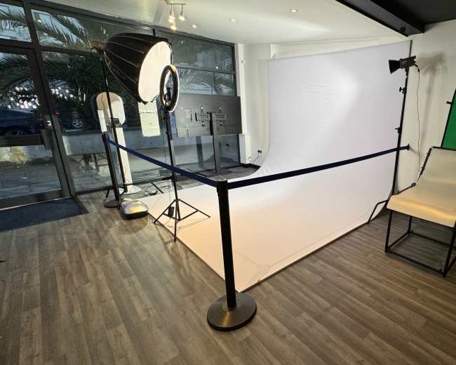 The image shows a photography studio setup with a white backdrop, a green screen, lighting equipment, and a folding chair.