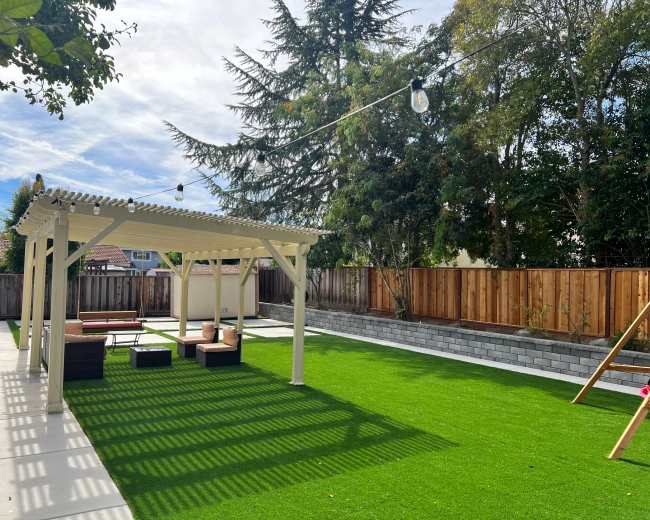 The image shows a landscaped backyard featuring a covered patio area, artificial grass, seating arrangements, and a wooden fence in the background.