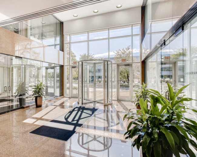 The image shows a modern entrance lobby with a revolving door, large glass windows, polished floors, and potted plants.