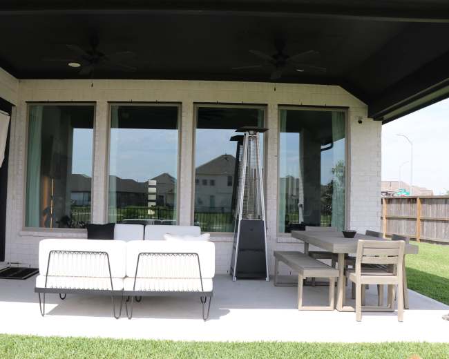 A covered patio featuring a white sofa, a dining table with chairs, and a tall patio heater next to large windows overlooking a lawn.